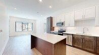 This well-appointed two-bedroom, one-bathroom unit at 3604 Rainbow Blvd offers a thoughtfully designed open-concept layout with modern finishes throughout. The combined kitchen and living area serves as the heart of the home, featuring a central island with a sleek quartz countertop, two-tone cabinetry pairing white shaker uppers with rich dark lower cabinets, full stainless steel appliances including a French door refrigerator, electric range, microwave, and dishwasher. White subway tile backsplash and recessed lighting complete the polished kitchen aesthetic. Light wood-look flooring flows throughout the main living areas, while large windows and a glass door bathe the living room in natural light and provide views of the surrounding community outdoor amenity spaces. The primary bedroom is generously sized, featuring soft carpet, elevated ceilings, and a stylish sliding barn door leading to the closet. The second bedroom also offers carpet flooring and a large window with community views. The bathroom is finished with large-format marble-look tile on both the floors and tub surround, a wood-toned vanity, and brushed nickel hardware. A dedicated laundry room includes a full-size top-loading washer and dryer with overhead shelving for added storage convenience.