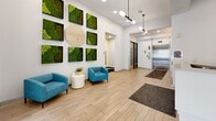 Located at 309 Oak St SE in Minneapolis, Minnesota, this thoughtfully designed single-floor amenity space offers a polished and modern environment for residents and visitors alike. The welcoming lobby immediately impresses with light wood-look tile flooring, soaring ceilings, and a striking living moss wall art installation bearing the Yugo branding — a vibrant, nature-inspired focal point that sets the tone for the entire space. Teal accent chairs and a small side table create an inviting seating nook, while an elevator and mailroom add practical convenience nearby. A sleek reception desk anchors a transitional hallway zone, providing a clean, professional atmosphere with easy access to adjacent areas. Floor-to-ceiling black-framed glass windows in the lounge area flood the space with abundant natural light and offer open views of the surrounding Minneapolis streetscape, complemented by sculptural pendant lighting and comfortable, stylishly upholstered seating. The kitchen is well-appointed with crisp white cabinetry, a teal subway tile backsplash, white quartz countertops, a matte black undermount sink, and stainless steel appliances. A colorful terrazzo-style floor and a casual round dining table with contemporary chairs make this kitchen equally functional and aesthetically pleasing — an ideal space for gathering and daily convenience.