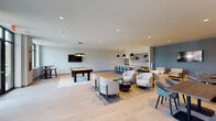 This contemporary amenity lounge is a well-appointed, open-plan space designed to accommodate a variety of social and recreational activities. Light wood-look tile flooring flows throughout, lending a clean, cohesive aesthetic that complements the neutral color palette of soft whites, warm creams, and calming blue-gray accent walls.

The space is organized into distinct functional zones. A game area anchors one side of the room, featuring a full-size pool table beneath a striking multi-shade pendant light fixture with gold interior accents. A wall-mounted flat-screen TV is positioned nearby, adding to the entertainment options. Adjacent to this is a cozy lounge zone with plush upholstered sofas and armchairs arranged around low coffee tables, ideal for casual conversation or relaxation. A decorative textured wall panel with sculptural accents and flanking wall sconces adds an artistic focal point to this seating area.

The kitchenette bar is outfitted with a large stainless steel refrigerator, an iridescent tile backsplash, open shelving with greenery, a sink, and a spacious counter with bar seating — perfect for hosting. A high-top table positioned beside a large window offers views of the surrounding outdoor greenery, filling the corner with natural light. Recessed ceiling lighting throughout ensures the entire space remains bright and welcoming at all hours.
