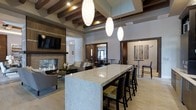 This single-floor clubhouse offers a thoughtfully designed, open-concept gathering space that balances elegance with comfort. The focal point of the main area is a generously sized granite island bar, surrounded by sleek high-back seating, making it an ideal spot for socializing or casual dining. Overhead, a striking coffered ceiling with rich, dark exposed wooden beams adds architectural depth, while a trio of elongated oval pendant lights cast a warm, inviting glow throughout the space.

Adjacent to the island, a comfortable lounge area centers around a modern stone-clad fireplace, accompanied by plush sectional seating and accented by a wall-mounted flat-screen television — perfect for relaxed gatherings. The neutral color palette of soft grays, warm wood tones, and stone textures creates a sophisticated yet welcoming ambiance throughout.

Further into the space, a secondary dining or café-style area features high-top tables and bar stools, ideal for smaller groups or informal meetings. Decorative artwork adds a refined touch to the walls. Natural light filters in through large windows and glass entry doors, brightening the interior and connecting the space to the outdoors. The overall design reflects a modern, upscale aesthetic suited for community events and everyday social interaction.