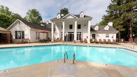 This amenity space combines an expansive outdoor pool area with a well-appointed interior, all anchored by a striking neoclassical-style clubhouse. The two-story white building serves as a dramatic backdrop to the pool, featuring prominent columned porticos, dormer windows, and French doors that open onto the pool deck. The generously sized swimming pool offers clear, blue water with convenient entry via stainless steel handrails, while the surrounding concrete deck provides ample space for relaxation. Brown lounge chairs line the pool's perimeter, and a shaded dining area with an orange market umbrella offers an alternative outdoor retreat. Mature trees frame the space, providing a natural, park-like setting. Inside the clubhouse, a compact but functional business center features wood-look flooring, built-in desks with computer workstations and a printer, and a bold teal-and-gold abstract wallcovering that creates a vibrant, modern contrast to the building's classic exterior. A windowed nook provides natural light to the workspace, while French glass-paned doors connect the interior rooms, maintaining an open, airy feel throughout.