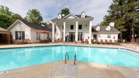 This amenity space combines an expansive outdoor pool area with a well-appointed interior, all anchored by a striking neoclassical-style clubhouse. The two-story white building serves as a dramatic backdrop to the pool, featuring prominent columned porticos, dormer windows, and French doors that open onto the pool deck. The generously sized swimming pool offers clear, blue water with convenient entry via stainless steel handrails, while the surrounding concrete deck provides ample space for relaxation. Brown lounge chairs line the pool's perimeter, and a shaded dining area with an orange market umbrella offers an alternative outdoor retreat. Mature trees frame the space, providing a natural, park-like setting. Inside the clubhouse, a compact but functional business center features wood-look flooring, built-in desks with computer workstations and a printer, and a bold teal-and-gold abstract wallcovering that creates a vibrant, modern contrast to the building's classic exterior. A windowed nook provides natural light to the workspace, while French glass-paned doors connect the interior rooms, maintaining an open, airy feel throughout.