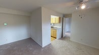 This single-floor residential unit offers a straightforward, functional layout designed for comfortable everyday living. The main living area is finished with neutral-toned carpet flooring and smooth white walls, creating a clean and understated aesthetic that provides a blank canvas for personalization. A partial dividing wall separates the living space from the kitchen, while an open pass-through cutout maintains a sense of visual connection between the two areas. The kitchen is equipped with warm-toned yellow wood cabinetry, a countertop with a built-in sink, and a microwave, all finished with light-toned flooring that contrasts gently with the carpeted living spaces. A hallway extends beyond the kitchen area, leading to additional rooms at the rear of the unit, suggesting a well-organized floor plan. The space benefits from consistent, even lighting throughout and maintains a bright, airy feel. The simple, no-frills design makes this unit highly adaptable for a range of uses, with its neutral palette and open sightlines contributing to an overall sense of openness and practicality.