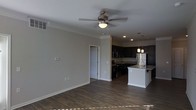 This single-story space showcases an open-concept layout that seamlessly connects the living area to the kitchen, creating a cohesive and functional floor plan. The living area features rich, dark hardwood-style flooring that extends throughout, complemented by neutral gray walls and crisp white baseboards and crown molding that add a polished, refined touch. A ceiling fan with a built-in light fixture is centrally positioned, providing both airflow and ambient lighting to the space.

The kitchen is a standout feature, designed with dark espresso cabinetry that offers ample storage and a bold, contemporary aesthetic. A central island with a light-toned countertop provides additional workspace and casual seating, anchored by stylish pendant lights hanging overhead. Stainless steel appliances, including a refrigerator and microwave, complete the kitchen's modern look. Recessed lighting illuminates the kitchen area efficiently.

A doorway visible from the living area suggests additional rooms beyond, contributing to the overall sense of a well-organized, practical layout. The combination of dark finishes against neutral walls creates a balanced, modern ambiance throughout the space.