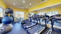 Located in Miami, FL, this fitness center is a well-equipped exercise room designed to support a full range of workout routines. The space is characterized by its vibrant yellow walls, white crown molding, and a ceiling fan with recessed lighting that keeps the environment bright and comfortable. Large, multi-pane windows line the walls, flooding the room with abundant natural light and offering picturesque views of a tranquil waterway and lush greenery — a refreshing backdrop for any workout session. Full-length mirrors are strategically placed to visually expand the space and assist with exercise form. The cardio section features multiple treadmills and an elliptical machine, while the strength training area includes a multi-station cable machine and various resistance equipment. A dedicated free weights station offers a tiered dumbbell rack alongside stability balls, catering to functional and flexibility training as well. The durable rubber flooring throughout provides safety and support. A wall-mounted television adds entertainment, and the overall layout makes efficient use of the compact footprint. The energetic color palette and natural surroundings create a motivating and inviting atmosphere for residents looking to maintain an active lifestyle.