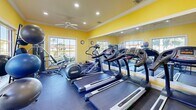 Located in Miami, FL, this fitness center is a well-equipped exercise room designed to support a full range of workout routines. The space is characterized by its vibrant yellow walls, white crown molding, and a ceiling fan with recessed lighting that keeps the environment bright and comfortable. Large, multi-pane windows line the walls, flooding the room with abundant natural light and offering picturesque views of a tranquil waterway and lush greenery — a refreshing backdrop for any workout session. Full-length mirrors are strategically placed to visually expand the space and assist with exercise form. The cardio section features multiple treadmills and an elliptical machine, while the strength training area includes a multi-station cable machine and various resistance equipment. A dedicated free weights station offers a tiered dumbbell rack alongside stability balls, catering to functional and flexibility training as well. The durable rubber flooring throughout provides safety and support. A wall-mounted television adds entertainment, and the overall layout makes efficient use of the compact footprint. The energetic color palette and natural surroundings create a motivating and inviting atmosphere for residents looking to maintain an active lifestyle.