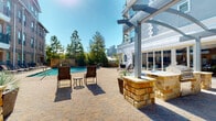 This outdoor amenity space is thoughtfully designed to offer a resort-like experience within an urban setting. At its centerpiece is a generously sized swimming pool with shimmering blue water, framed by a wide paver deck finished in a stamped brick pattern that lends warmth and texture throughout the space. Rows of lounge chairs line the poolside, providing ample space for sunbathing, while mature trees and a wrought iron perimeter fence create a sense of privacy without obstructing the surrounding city skyline views.

Adjacent to the pool, a covered pergola structure with steel arched framing defines an outdoor living and dining zone. This area features a built-in outdoor kitchen constructed with natural stacked stone, complete with a gas grill and counter space, alongside wicker dining furniture suited for group gatherings. Decorative planters with ornamental grasses add greenery and visual interest throughout the patio.

A separate seating area near a natural stone bar counter offers a more intimate setting for relaxing. The overall design blends natural stone masonry with contemporary architectural elements, creating a cohesive and inviting outdoor environment that benefits from abundant natural sunlight throughout the day.