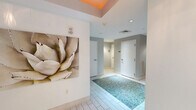 Located at 700 SW 78th Ave in Plantation, FL, this well-appointed hallway serves as a transitional and welcoming corridor space. The design immediately draws attention with a bold orange-painted tray ceiling detail near the entrance, adding a vibrant pop of color against the otherwise neutral white and soft gray palette throughout. Large-format white tile flooring runs the length of the hallway, punctuated by a striking inset section of turquoise pebble mosaic tile that serves as a distinctive decorative focal point underfoot. The walls are adorned with curated artwork, including a large-scale botanical canvas and colorful abstract framed pieces, lending the space an artistic, gallery-like quality. Recessed lighting provides clean, even illumination throughout. A set of glass-paneled entry doors at one end opens to what appears to be a fitness facility, allowing natural light to filter into the corridor. Additional doors along the hallway provide access to restrooms, including an ADA-accessible women's restroom, as well as a mechanical room. A ventilation grille is neatly integrated into one wall. The overall ambiance is clean, modern, and thoughtfully designed, balancing functionality with aesthetic appeal to create an inviting passage for building occupants and visitors alike.