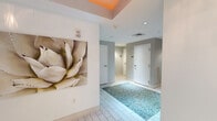Located at 700 SW 78th Ave in Plantation, FL, this well-appointed hallway serves as a transitional and welcoming corridor space. The design immediately draws attention with a bold orange-painted tray ceiling detail near the entrance, adding a vibrant pop of color against the otherwise neutral white and soft gray palette throughout. Large-format white tile flooring runs the length of the hallway, punctuated by a striking inset section of turquoise pebble mosaic tile that serves as a distinctive decorative focal point underfoot. The walls are adorned with curated artwork, including a large-scale botanical canvas and colorful abstract framed pieces, lending the space an artistic, gallery-like quality. Recessed lighting provides clean, even illumination throughout. A set of glass-paneled entry doors at one end opens to what appears to be a fitness facility, allowing natural light to filter into the corridor. Additional doors along the hallway provide access to restrooms, including an ADA-accessible women's restroom, as well as a mechanical room. A ventilation grille is neatly integrated into one wall. The overall ambiance is clean, modern, and thoughtfully designed, balancing functionality with aesthetic appeal to create an inviting passage for building occupants and visitors alike.