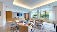 Located at 695 Atlantic St in Stamford, Connecticut, this elegantly appointed lounge space offers a refined and welcoming atmosphere designed for comfort and style. The room is defined by a striking tray ceiling fitted with warm indirect LED lighting, casting a soft ambient glow across the interior. Large windows dressed with sheer curtains flood the space with natural light while framing views of surrounding greenery, creating a seamless connection between the interior and the outdoors. The flooring transitions between large-format tile and a patterned area rug, anchoring the central seating arrangement. The furnishings include a curated mix of upholstered armchairs in neutral tones, a tufted cognac leather bench, and a tiered circular wood coffee table, all arranged to encourage conversation and relaxation. A textured mosaic accent wall serves as a bold focal point, complemented by a wall-mounted flat-screen television and warm wood cabinetry with built-in storage below. Vertical wood slat partitioning adds architectural depth and visual interest near the entry area. A navy upholstered banquette with a bar-height table provides an additional seating option. The overall design palette — soft blues, warm wood tones, and neutral textiles — conveys a polished, contemporary aesthetic suited for a communal or hospitality setting.