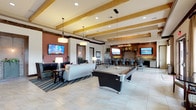 This expansive single-floor clubhouse lounge is thoughtfully designed to accommodate a variety of social and recreational activities. The space is anchored by an open-plan layout with large-format tile flooring throughout, exposed wood ceiling beams, and recessed lighting complemented by pendant fixtures that add warmth and character to the interior.

The main lounge area features comfortable seating arrangements, including a sofa and accent chairs positioned around a striped area rug, with wall-mounted flat-screen televisions providing entertainment. A full-size billiards table serves as a central focal point, making this zone ideal for casual gatherings.

Adjacent to the lounge is a bar-height dining area anchored by a dramatic stone fireplace surround and display shelving. Dark cabinetry lines the wall beneath multiple mounted screens, and a long communal high-top table with sleek metal chairs provides ample seating for group dining or socializing. French doors in this area open directly to an outdoor patio, flooding the space with natural light.

A dedicated game room alcove houses a shuffleboard table and benefits from its own pendant lighting and window views. A decorative river rock inlay in the flooring adds a distinctive natural design element that runs through portions of the space.

Restroom access and additional seating areas round out the functionality of this well-appointed amenity space.