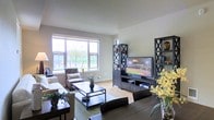 This single-floor space offers a thoughtfully designed living area that balances comfort and style. The living room serves as the heart of the home, bathed in natural light streaming through large windows that frame views of lush greenery outside. Light hardwood flooring runs throughout, complementing the neutral, soft-toned walls and creating a warm, inviting atmosphere.

The living area is anchored by a bold, dark-finished entertainment unit, complete with matching shelving towers featuring decorative X-cross detailing — a standout architectural accent that adds character to the space. A large flat-screen television is centrally positioned, flanked by open shelving that provides both display and storage opportunities.

Comfortable seating surrounds a sleek, dark metal-framed coffee table, bringing together a cohesive and functional arrangement. A patterned accent chair positioned near the window offers a cozy reading nook with an outdoor view. Touches of greenery and floral arrangements throughout the room add a fresh, lively energy to the overall ambiance.

The space is well-proportioned, offering an open and airy feel without feeling oversized. Natural light, clean lines, and a balanced mix of dark and light tones make this a welcoming and visually harmonious living environment.