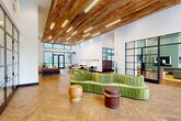 Located at 611 O'Keefe Ave in New Orleans, Louisiana, this expansive single-floor lobby and hallway space offers a thoughtfully designed environment that blends modern aesthetics with warm, organic materials. Spanning a generous footprint, the space immediately impresses with its soaring double-height ceilings partially clad in reclaimed wood planking, illuminated by sleek linear pendant lighting that casts a warm, inviting glow throughout. The flooring features a refined herringbone-patterned wood tile that anchors the space with understated elegance.

Floor-to-ceiling black-framed glass walls and partitions divide the interior into distinct zones while preserving an open, airy feel. Large street-facing windows flood the entrance with abundant natural light, framing views of the surrounding urban streetscape. Interior greenery, including tall fiddle-leaf fig trees and potted plants, adds life and texture throughout.

The lobby area is furnished with a distinctive curved green velvet sofa and sculptural wooden stools, creating a welcoming lounge focal point. Adjacent areas offer collaborative workspaces with long communal tables and seating, a dedicated lounge with softer furnishings and curated artwork, and a reception desk with dark fluted paneling. Steel-framed glass partition walls enclose additional rooms visible from the main hall, ensuring transparency and connectivity throughout the entire space.