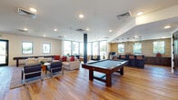 This two-floor amenity facility offers a well-appointed and thoughtfully designed environment ideal for socializing and recreation. The ground floor welcomes visitors through a bright, open lobby featuring a mix of large-format tile and warm hardwood flooring, with glass doors that frame views of an outdoor terrace and pool area beyond.

The main lounge and game room is a standout space, anchored by rich hardwood floors and filled with natural light from floor-to-ceiling windows along the perimeter. A full-size billiard table commands the center, complemented by a shuffleboard table, multiple seating areas with plush sofas and accent chairs, and a bar and kitchenette with textured stone tile backsplash. A secondary kitchen offers dark wood cabinetry, granite countertops, stainless steel appliances, and a striking mosaic tile feature wall with dual mounted flat-screen televisions.

The upper level presents a sophisticated lounge with soaring double-height windows and sliding glass doors that open to a furnished rooftop terrace. The terrace features teak deck tiles, outdoor seating, a dining table, and a built-in grill station, offering panoramic views of the surrounding area. A dedicated kitchenette with dark cabinetry, a full-size refrigerator, and generous counter space rounds out the upper floor's amenities.
