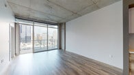 This contemporary high-rise unit showcases an industrial-modern aesthetic defined by exposed concrete ceilings, wood-look plank flooring, and an abundance of natural light. The open living area is anchored by dramatic floor-to-ceiling glass walls and sliding doors that frame sweeping panoramic views of a vibrant downtown skyline, creating a seamless connection between the interior and the urban landscape beyond. Exposed ductwork and a ceiling fan add to the raw, loft-style character of the space.

The kitchen is well-appointed with stainless steel appliances, white upper cabinetry, wood-toned lower cabinets, a subway tile backsplash, and a generous island with a white countertop that provides both prep space and casual seating. Track lighting and a statement pendant lamp illuminate the kitchen area.

The bathroom features large-format tile flooring, a glass-enclosed shower, and a dark wood vanity with a clean white countertop, offering a refined, spa-like finish.

A private balcony accessed via sliding glass doors offers an open-air vantage point with glass railings, providing unobstructed views of the surrounding cityscape. The overall design balances industrial elements with modern finishes throughout.