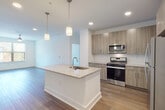 This single-floor residence showcases a contemporary open-concept design that seamlessly connects the kitchen and living areas. The kitchen serves as the centerpiece of the space, anchored by a substantial white island topped with speckled granite countertops and an undermount sink. Two pendant lights hang above the island, providing focused task lighting alongside recessed ceiling fixtures. Light wood-toned cabinetry lines the walls, paired with stainless steel appliances including a gas range, microwave, and refrigerator. Wood-look plank flooring extends throughout the main living areas, adding warmth and continuity.

The living area benefits from natural light provided by large windows with outdoor views of surrounding greenery. Bedrooms are finished with neutral-toned carpet and feature windows that bring in ample natural light. The primary bathroom is well-appointed with a dual-sink granite vanity, wood-toned cabinetry with drawers, a large framed mirror, a walk-in shower, and direct access to a walk-in closet with built-in shelving. A secondary bathroom features a similarly styled granite vanity with an undermount sink and a full-wall mirror. The overall aesthetic is clean, modern, and cohesive throughout.