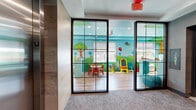 Located on the third floor at 975 Bonnel Ct in Union, New Jersey, this thoughtfully designed space combines a welcoming lobby corridor with a lively, dedicated children's play area. The lobby entrance features textured gray wall coverings, recessed lighting, elevator access, and stairwell entry, accented by a decorative wood-framed credenza topped with colorful ceramic vases and a large round mirror — lending a polished, contemporary feel.

Accessible through a striking floor-to-ceiling glass partition with a dark metal frame, the children's play area is a fully immersive environment. The walls are adorned with vibrant, hand-painted cartoon murals depicting trees, animals, and whimsical landscapes that wrap the entire room, complemented by a painted teal ceiling that enhances the playful atmosphere. Natural light filters in through windows overlooking the surrounding neighborhood.

The play area is furnished with age-appropriate features including built-in cubbies and shelving stocked with toys and books, a central play structure with open compartments for imaginative play, child-sized tables and chairs, and a chalkboard easel. Light hardwood flooring runs throughout the play space, offering a warm contrast to the vivid surroundings. Together, these elements create an engaging, safe, and stimulating environment well-suited for young children.