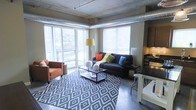 This single-floor space showcases a distinctive industrial-modern aesthetic defined by its exposed concrete ceiling and visible silver ductwork running along the perimeter — hallmark features of loft-style architecture. The living area is generously proportioned, anchored by large floor-to-ceiling windows and a sliding glass door that floods the interior with natural light and offers views of an outdoor balcony and surrounding greenery beyond. The polished concrete flooring adds to the industrial character while maintaining a sleek, contemporary feel. A bold geometric-patterned area rug delineates the main seating zone, which is centered around a dark navy sofa accented with colorful throw pillows and complemented by a warm-toned accent chair. A glass-top coffee table adds a lighter visual element to the arrangement. To one side, a compact workspace or dining area with a high-top table and stool provides functional versatility. Built-in shelving or cabinetry in a dark finish is visible along one wall, offering storage and display space. The overall ambiance balances raw industrial elements with warm, curated furnishings, creating a comfortable and visually dynamic living environment.