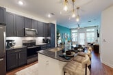 This thoughtfully designed single-floor residence combines modern aesthetics with functional living spaces. The open-concept kitchen is a standout centerpiece, featuring dark navy shaker-style cabinetry with brushed gold hardware, rich dark granite countertops, and a herringbone tile backsplash. Stainless steel appliances, including a French door refrigerator, over-the-range microwave, and electric range, provide a fully equipped cooking experience. A kitchen island with an undermount sink and seating for multiple bar stools doubles as a dining and prep space, illuminated by elegant gold pendant lights overhead. The kitchen flows seamlessly into the living area, which is anchored by a bold teal accent wall and warm hardwood-style flooring, creating a vibrant yet inviting atmosphere. Natural light pours in through multiple windows and glass doors, enhancing the open feel. The bedroom is generously proportioned, featuring soft carpeting, a ceiling fan, a matching teal accent wall, and ample natural light. The bathroom offers a soaking tub and shower combination with neutral tile surround and built-in open shelving for organized storage. Throughout the space, consistent use of gold accents, teal tones, and contrasting dark and light finishes creates a cohesive, contemporary design identity.