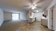This single-floor residence offers an open-concept layout that seamlessly connects the kitchen and living areas, creating a cohesive and functional living environment. The kitchen is outfitted with white cabinetry, a built-in sink, a dishwasher, a range, and a refrigerator, complemented by tile flooring and a skylight that introduces natural light overhead. The countertops provide practical workspace, and under-cabinet storage ensures efficient use of space. Transitioning into the living area, the flooring shifts to wall-to-wall carpeting, lending a comfortable and warm atmosphere. A ceiling fan with a light fixture is centrally positioned, providing both airflow and ambient lighting throughout the shared space. A large sliding glass door along the far wall floods the living area with natural light and offers direct access to an exterior patio or balcony, framing views of the outdoors. The textured ceilings are consistent throughout, and a hallway visible from the main area leads to additional rooms. The open flow between the kitchen and living space makes this layout well-suited for everyday living and casual entertaining.