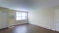 This single-floor space offers a straightforward and functional layout designed with simplicity in mind. The main living area is characterized by smooth hardwood flooring in a warm, medium-toned finish that extends throughout the room, providing a clean and cohesive look underfoot. The walls are finished in a soft, neutral beige tone that reflects light evenly and contributes to a calm, understated ambiance. A window along one wall allows natural light to filter into the space, brightening the interior and creating an open, airy feel. A baseboard heating system runs along the base of the walls, ensuring efficient and consistent warmth throughout. An additional wall-mounted unit, visible near the window, provides supplementary climate control. The high, smooth ceiling adds to the sense of openness within the room. Clean white baseboards frame the flooring neatly, adding a crisp contrast to the neutral walls. The overall design is minimal and move-in ready, with no excess ornamentation, making it a versatile space easily adaptable to a variety of furnishing styles and personal preferences. The uncluttered layout allows for flexible furniture arrangements to suit different needs.