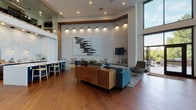 The Alexan Marymoor clubhouse in Redmond, Washington offers a thoughtfully designed communal space that blends modern architecture with warm, inviting interiors. Rich hardwood flooring runs throughout, complementing the soaring double-height ceilings that lend an airy, open ambiance to the entire floor.

At the heart of the space is a generously proportioned lounge area featuring curated seating arrangements, including plush blue sofas and accent chairs, anchored by a mid-century modern credenza and tasteful artwork. Recessed and track lighting highlight architectural details and art pieces, adding depth to the crisp white walls.

The kitchenette bar area is a standout feature, showcasing a bold teal island with a white quartz countertop, woven bar stools, pendant lighting, and a stone-tiled backsplash — all complementing a professional-grade coffee and beverage station.

A dedicated reading nook lined with wall-mounted magazine displays and a sunken lounge area with textured tile walls provide intimate retreat spaces. Floor-to-ceiling black-framed glass walls and doors flood the interior with natural light and open directly onto a spacious outdoor terrace. The terrace is surrounded by lush Pacific Northwest tree canopy, offering a serene backdrop for outdoor seating and dining.