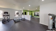 This single-story fitness center is thoughtfully designed to accommodate a range of workout activities across multiple interconnected zones. The main cardio area features rubber flooring throughout and houses a selection of equipment including treadmills, a stationary bike, and an elliptical machine, all positioned to allow comfortable spacing between users. Recessed lighting and a ceiling fan provide adequate illumination and air circulation across the open workout floor.

A dedicated strength training area is distinguished by vibrant green accent walls and contains a weight bench, a rack of dumbbells, medicine balls, and a stability ball. A large wall-mounted mirror in this zone enhances the sense of space while allowing users to monitor their form. Windows throughout the facility bring in natural light and provide views of the surrounding exterior.

The layout flows between zones through an open corridor, with a glass-paneled entry door visible at one end offering access to the outdoors. Restroom facilities with separate entrances are available on-site, adding convenience for users. The overall design balances functionality with a clean, modern aesthetic suited to a community fitness environment.