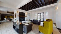 Located at 2785 Polo Club Blvd in Lexington, Kentucky, this single-story community clubhouse offers a well-appointed gathering space designed with a contemporary aesthetic and high-end finishes throughout. The main lounge serves as the heart of the facility, featuring a striking fireplace with a rustic wood mantle and dark herringbone tile surround, flanked by built-in shelving and anchored by exposed dark wood beam ceiling details. Tufted charcoal sofas, olive accent chairs, and patterned area rugs create an inviting, sophisticated atmosphere, while large French doors and transom windows flood the space with natural light. Adjacent to the lounge, the entertainer's kitchen features expansive white quartz island countertops, tufted bar seating, sleek pendant lighting, and stainless steel appliances beneath a distinctive textured bronze ceiling. The herringbone-patterned tile flooring extends throughout this area, which also accommodates a billiards table, making it ideal for social gatherings. A dedicated media room offers plush carpeting, bold plum accent walls, a large wall-mounted flat-screen television, and comfortable seating for an intimate viewing experience. A separate lounge nook with bold yellow chairs and ample natural light provides additional casual seating. Together, these spaces deliver a cohesive, upscale environment designed for community engagement and relaxation.