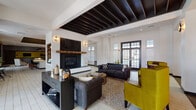 Located at 2785 Polo Club Blvd in Lexington, Kentucky, this single-story community clubhouse offers a well-appointed gathering space designed with a contemporary aesthetic and high-end finishes throughout. The main lounge serves as the heart of the facility, featuring a striking fireplace with a rustic wood mantle and dark herringbone tile surround, flanked by built-in shelving and anchored by exposed dark wood beam ceiling details. Tufted charcoal sofas, olive accent chairs, and patterned area rugs create an inviting, sophisticated atmosphere, while large French doors and transom windows flood the space with natural light. Adjacent to the lounge, the entertainer's kitchen features expansive white quartz island countertops, tufted bar seating, sleek pendant lighting, and stainless steel appliances beneath a distinctive textured bronze ceiling. The herringbone-patterned tile flooring extends throughout this area, which also accommodates a billiards table, making it ideal for social gatherings. A dedicated media room offers plush carpeting, bold plum accent walls, a large wall-mounted flat-screen television, and comfortable seating for an intimate viewing experience. A separate lounge nook with bold yellow chairs and ample natural light provides additional casual seating. Together, these spaces deliver a cohesive, upscale environment designed for community engagement and relaxation.
