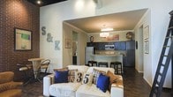 This single-floor space showcases a thoughtfully designed open-concept layout that blends industrial and contemporary aesthetics. The living area anchors the space with a plush white sofa adorned with decorative pillows in navy blue and Union Jack prints, reflecting a bold, eclectic personality. Exposed brick walls add a raw, industrial character, while the dark hardwood flooring grounds the space with warmth and sophistication. Notably high ceilings with visible ductwork reinforce the industrial-chic design language throughout.

The kitchen is seamlessly integrated into the open floor plan, featuring dark cabinetry, a tiled backsplash, stainless steel appliances — including a refrigerator and microwave — and a peninsula-style breakfast bar with bar stools, making it ideal for casual dining and entertaining. A pendant light fixture above the bar area provides focused illumination, complementing the overall warm lighting scheme.

Decorative elements, including large metal letters mounted on the brick wall and framed artwork, add a personalized and curated touch to the space. A bistro-style seating nook is visible near the brick wall, offering a cozy retreat. The overall ambiance is modern and urban, with a welcoming and lived-in character that balances style with comfort.