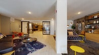 This thoughtfully designed single-floor amenity space offers a dynamic and versatile environment suited for both relaxed gatherings and active socializing. The layout is organized into several distinct zones, each with its own character while maintaining a cohesive, contemporary aesthetic throughout.

The main lounge area features large floor-to-ceiling windows dressed with sheer white curtains that flood the space with abundant natural light. Plush seating, including a tufted velvet sofa and rich leather sectional, is complemented by patterned area rugs and accent tables in bold yellow, creating a warm and inviting atmosphere.

A stylish wet bar anchors one section of the space, complete with a sleek dark island topped with a white surface, wood-stool seating, and elegant teardrop pendant lighting overhead. Textured wood-paneled walls and a grid of decorative black wall art add visual depth and sophistication to this area.

The recreation zone is well-appointed with a pool table, shuffleboard, arcade games, and communal high-top tables surrounded by colorful accent chairs — all illuminated by a modern linear pendant fixture. Built-in shelving with warm brass accents and curated décor further elevate the lounge aesthetic. Light porcelain tile flooring runs seamlessly throughout, tying the entire space together with a clean, polished finish.