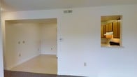 This single-story space presents a functional, open layout characterized by clean white walls and a neutral color palette throughout. The main living area features carpeted flooring, transitioning to a smoother, light-toned floor surface in the adjoining room visible through a wide open doorway. This secondary room, likely a dining area or additional living space, benefits from its own ceiling vent and wall outlets, suggesting versatile use potential.

A pass-through opening on the right side of the main wall offers a direct sightline into the kitchen, where wooden cabinetry in a warm brown finish and light countertops are visible. This architectural detail creates a sense of connectivity between the living and kitchen areas while maintaining defined zones within the space.

Central to the main wall is a wall-mounted thermostat, indicating a climate control system. A ceiling-mounted HVAC vent ensures consistent air distribution throughout. The overall design is straightforward and practical, with white baseboards framing the floors and standard electrical outlets positioned along the walls. The space offers a clean, move-in-ready canvas with a simple, utilitarian aesthetic.