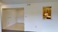 This single-story space presents a functional, open layout characterized by clean white walls and a neutral color palette throughout. The main living area features carpeted flooring, transitioning to a smoother, light-toned floor surface in the adjoining room visible through a wide open doorway. This secondary room, likely a dining area or additional living space, benefits from its own ceiling vent and wall outlets, suggesting versatile use potential.

A pass-through opening on the right side of the main wall offers a direct sightline into the kitchen, where wooden cabinetry in a warm brown finish and light countertops are visible. This architectural detail creates a sense of connectivity between the living and kitchen areas while maintaining defined zones within the space.

Central to the main wall is a wall-mounted thermostat, indicating a climate control system. A ceiling-mounted HVAC vent ensures consistent air distribution throughout. The overall design is straightforward and practical, with white baseboards framing the floors and standard electrical outlets positioned along the walls. The space offers a clean, move-in-ready canvas with a simple, utilitarian aesthetic.