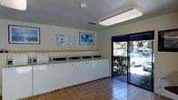 This single-story laundry facility is designed for straightforward, communal use. The main room features a continuous row of top-loading washing machines lined along one wall, providing ample capacity for multiple users. The machines are accompanied by an overhead shelf, offering a convenient surface for laundry supplies and folded items.

The room is finished with warm terra cotta ceramic tile flooring, which is both durable and easy to maintain. Overhead, a long fluorescent light fixture provides bright, even illumination throughout the space. The ceiling features a textured finish and includes a centrally mounted smoke detector, along with a ventilation register for climate control.

Natural light enters through a large glass sliding door positioned along one wall, fitted with a dark privacy valance at the top. This door also provides a view of a lush, green landscaped area outside, adding a pleasant visual connection to the outdoors.

Informational signage, including washing instructions, is posted on the wall alongside a framed decorative print, contributing a modest sense of order and character to the functional space. Overall, the facility is clean, well-lit, and practical.