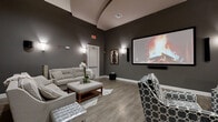 Located in Winter Garden, Florida, this sophisticated screening and entertainment lounge offers a well-appointed single-floor layout designed for an immersive cinematic experience. The room is anchored by a large wall-mounted projection screen paired with a soundbar, making it an ideal venue for movie nights or private gatherings. Tall ceilings with a subtle tray detail lend a sense of vertical openness to the space, while elegant wall sconces cast warm, ambient lighting throughout the charcoal-toned interior. The rich, dark gray walls create a dramatic, theater-like atmosphere that complements the wood-look plank flooring underfoot. Seating is plentiful and varied, with a sectional sofa, plush armchairs in a bold geometric pattern, and a tufted ottoman serving as a central coffee table — all arranged to maximize sightlines to the screen. A cowhide rug adds a sophisticated, textural accent to the seating area. Along one wall, a built-in wet bar features warm cherry wood cabinetry, ample upper and lower storage, and a sweeping granite countertop with an integrated sink — ideal for entertaining. Decorative artwork and curated accessories complete the polished, upscale aesthetic of this inviting entertainment space.