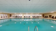 Located at 4946 Snapfinger Woods Dr in Decatur, Georgia, this expansive single-floor indoor aquatic facility offers a generously sized, climate-controlled environment ideal for recreation and leisure. The centerpiece of the space is a large rectangular lap pool with vivid turquoise water, illuminated from below by underwater lighting that enhances its visual appeal. Stainless steel entry railings provide safe and convenient pool access.

Surrounding the pool is a broad, textured deck finished in a durable aggregate surface, providing ample space for circulation and relaxation. Along one entire side of the pool, a row of woven strap lounge chairs lines the perimeter, positioned in front of a continuous series of white-framed windows and glass doors that allow natural light to filter in and offer views of the exterior grounds.

A raised seating area bordered by a natural stone wall and metal railings serves as a distinct lounge zone, furnished with upholstered sofas and side tables, creating a comfortable retreat overlooking the pool. Artwork is displayed on the walls, adding a decorative touch to the otherwise utilitarian setting. The drop-panel ceiling is fitted with recessed and flush-mount lighting throughout, ensuring consistent illumination across the entire facility. Multiple exit doors provide convenient access to the outdoor areas.