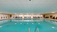 Located at 4946 Snapfinger Woods Dr in Decatur, Georgia, this expansive single-floor indoor aquatic facility offers a generously sized, climate-controlled environment ideal for recreation and leisure. The centerpiece of the space is a large rectangular lap pool with vivid turquoise water, illuminated from below by underwater lighting that enhances its visual appeal. Stainless steel entry railings provide safe and convenient pool access.

Surrounding the pool is a broad, textured deck finished in a durable aggregate surface, providing ample space for circulation and relaxation. Along one entire side of the pool, a row of woven strap lounge chairs lines the perimeter, positioned in front of a continuous series of white-framed windows and glass doors that allow natural light to filter in and offer views of the exterior grounds.

A raised seating area bordered by a natural stone wall and metal railings serves as a distinct lounge zone, furnished with upholstered sofas and side tables, creating a comfortable retreat overlooking the pool. Artwork is displayed on the walls, adding a decorative touch to the otherwise utilitarian setting. The drop-panel ceiling is fitted with recessed and flush-mount lighting throughout, ensuring consistent illumination across the entire facility. Multiple exit doors provide convenient access to the outdoor areas.