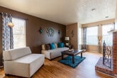 This single-floor residence offers a thoughtfully designed layout that balances comfort and functionality throughout. The living room serves as the heart of the home, featuring warm hardwood-style flooring, a textured ceiling, and a bold dark accent wall that anchors the space. A striking brick fireplace connects the living and dining areas, adding character and warmth. Multiple windows allow natural light to fill the room. Adjacent to the living area, the dining space accommodates a raised table and seating, with recessed lighting overhead. The kitchen is equipped with warm-toned wood cabinetry, dark countertops, black appliances including a range, microwave, and refrigerator, and direct access to a dedicated pantry for added storage. The two bedrooms are carpeted and comfortably sized, each featuring ceiling fans and windows. One bedroom includes a spacious walk-in closet with built-in shelving, providing ample storage. The full bathroom features warm wood vanity cabinetry, a wide countertop with an integrated sink, a large mirror, and hardwood-style flooring. Overall, the home presents a cohesive and livable layout with practical finishes throughout.
