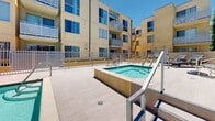 Located at 9850 Zelzah Ave in the Northridge neighborhood of Los Angeles, this outdoor amenity area offers a well-appointed communal pool and spa experience under the abundant Southern California sun. The centerpiece of the space is a generously sized rectangular swimming pool with clear turquoise water, accessible via chrome handrails and surrounded by a broad concrete pool deck. Adjacent to the main pool sits an elevated spa, finished with tiled surrounds and its own step access, providing a distinct relaxation zone within the same open-air environment. The deck is finished with large-format light stone tiles, lending a clean and modern aesthetic throughout the courtyard. Lounge chairs are positioned along the perimeter, offering comfortable spots to enjoy the open sky and natural light. The entire area is enclosed by sleek vertical metal fencing with a gated entry, ensuring both security and a sense of privacy. The warm-toned stucco exterior walls of the surrounding multi-story building frame the space on multiple sides, creating a sheltered and semi-private atmosphere. Mature trees are visible beyond the perimeter wall, adding a touch of greenery to the urban setting. This outdoor space is designed to serve as a resort-style retreat within a vibrant Los Angeles community.