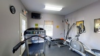 The fitness room at Claysville Landing is a single-floor space designed to support residents' exercise needs in a functional, well-organized environment. The room features light gray walls and durable dark flooring suited for high-traffic workout use. Natural light enters through two windows dressed with colorful striped valances, contributing to a bright and energizing atmosphere. A wall-mounted flat-screen television is positioned for easy viewing during workouts. The equipment selection includes a treadmill, a multi-function cable weight machine, and an elliptical trainer, providing options for both cardiovascular and strength training exercises. Overhead fluorescent lighting ensures the space remains well-lit throughout the day. Informational notices are posted on the walls, keeping users informed of guidelines and instructions. The layout makes efficient use of the available space, positioning equipment to allow reasonable movement between machines. Overall, the fitness room at Claysville Landing offers a practical and accessible amenity for residents looking to maintain an active lifestyle without leaving the community.