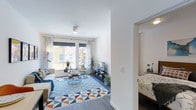 This single-floor space is thoughtfully designed to maximize comfort and natural light. The living room serves as the heart of the home, featuring large French-style windows dressed with patterned curtains and roller shades that flood the room with daylight while offering views of the surrounding exterior. Clean white walls and light-toned ceilings create an airy, open atmosphere throughout.

The living area is anchored by a bold geometric-patterned area rug in shades of blue, orange, and white, which adds a vibrant, contemporary energy to the space. A comfortable white sofa and coordinating armchair are arranged around a round metal coffee table, creating an inviting seating area. A media console with a flat-screen television and an assortment of indoor plants add both function and a natural, lived-in warmth. Dark hardwood-style flooring runs throughout, providing a rich contrast to the bright walls.

Through an open doorway, the bedroom is visible and carries the same clean, modern aesthetic, with soft textiles and warm ambient lighting. The layout promotes a natural flow between living and sleeping areas, making the space feel cohesive and well-connected. Overall, the design balances a modern sensibility with warm, welcoming details.