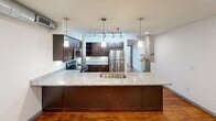 Located at 1101 Hawthorne Lane in Charlotte, NC, this two-story loft showcases an inspired mix of industrial heritage and contemporary design. The main level features a well-appointed kitchen with granite countertops, dark cabinetry, stainless steel appliances, a large island with a double sink, and track lighting. Rich hardwood floors run throughout, complemented by exposed wood beam ceilings, original structural columns, and soaring ceiling heights that create a dramatic, open ambiance. The bedroom offers built-in closet storage, while both bathrooms feature granite vanities and clean, modern tile finishes. The upper loft level adds versatile additional living space, accessible via an interior staircase.