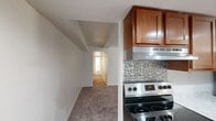 This single-floor, 2-bedroom, 1-bathroom residence offers a well-organized layout across a modest footprint. The galley-style kitchen is a functional highlight, featuring warm medium-toned wood cabinetry, marble-style countertops, a stainless steel refrigerator, dishwasher, and range, complemented by recessed lighting and light hardwood-style flooring. A decorative patterned tile backsplash adds a distinct visual element near the range area. The open connection between the kitchen and the carpeted main living area creates a natural flow through the unit.

Both bedrooms are carpeted and bathed in natural light through horizontal-blind-fitted windows that offer outdoor views of surrounding greenery. The primary bedroom includes a closet with a rod system, while the second bedroom also features its own closet, providing practical storage in each room. The hallway connects the bedrooms to the shared bathroom, which includes a white vanity with under-cabinet storage, a mirrored medicine cabinet, and light hardwood-style flooring. The neutral color palette throughout — white walls, beige carpet, and clean trim — contributes to a bright, cohesive ambiance across the entire unit.