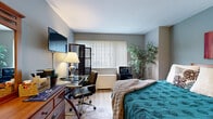 Located at 12540 Edgewater Drive in Lakewood, Ohio, this compact yet efficiently designed one-bedroom, one-bathroom unit makes smart use of its space across a single floor. The main living area serves as both a sleeping and functional workspace, highlighted by warm light wood flooring that runs throughout and cool-toned blue-gray walls that lend a calm, cohesive ambiance.

The generously sized bedroom accommodates a queen bed alongside a dedicated work area featuring a glass-top desk, ergonomic chair, and a wall-mounted flat-screen TV. A dark wood folding screen partition adds a stylish, flexible element to the room's layout. Natural light enters through a large window dressed with vertical blinds, brightening the space throughout the day. A tall indoor plant and tasteful wall art, including a bold red and neutral canvas piece, add personality and warmth to the room. A wood dresser with mirror provides additional storage.

The bathroom is clean and functional, featuring a full bathtub with shower and a crisp white surround, complemented by light flooring and neutral walls. The overall design of this unit prioritizes comfort and practicality, making it a well-suited space for individuals seeking an efficient, move-in-ready home in the Lakewood area.