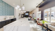 This well-appointed single-floor space offers a generously sized open-plan layout integrating a kitchen, dining area, and living room in one cohesive environment. The design aesthetic is modern and sophisticated, characterized by soaring ceilings that amplify the sense of volume and an abundance of natural light streaming through large floor-to-ceiling windows and glass doors, offering views of the surrounding outdoor area.

The kitchen features sleek dark cabinetry topped with a striking white marble-veined countertop that extends into a substantial island. A bold teal grid-paneled accent wall serves as a dramatic backdrop above the counters, adding a distinctive architectural focal point. Recessed lighting and globe pendant fixtures with brass hardware provide layered illumination throughout.

The living area centers around a floor-to-ceiling textured tile fireplace surround with a mounted television and integrated log storage. Thoughtfully arranged seating, patterned area rugs, and curated artwork contribute warmth and character. A dedicated dining nook features a dark table alongside large glazed walls, maximizing outdoor views. The flooring transitions between large-format tile and wood-look planks, delineating zones while maintaining visual continuity.