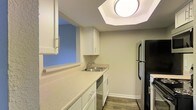 This single-floor space offers a clean, functional layout designed with practicality and comfort in mind. The kitchen is a standout feature, showcasing crisp white cabinetry that extends to the upper walls, providing ample storage for all culinary essentials. The countertops offer generous prep space and are finished in a neutral tone that complements the overall color palette of the space. A stainless steel double-basin sink sits beneath a pass-through window that opens to an adjacent area, allowing natural light to filter through and creating a sense of openness. Black appliances, including a full-sized refrigerator and stove, add a bold contrast against the white cabinetry, while a built-in dishwasher contributes to the kitchen's practical design. The flooring throughout features wood-look planks that lend warmth and durability underfoot. A large flush-mount ceiling light provides bright, even illumination across the space. The walls are finished in soft, neutral tones, contributing to a calm and inviting ambiance. Overall, the space is thoughtfully designed to maximize functionality while maintaining a tidy, modern aesthetic suited for everyday living.