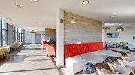 Located at 5104 Jacobs Rd in Seneca, SC, this single-floor community living space offers a generously sized, open-plan layout designed for comfort and social interaction. The space is defined by a striking combination of modern architecture and warm, inviting design elements. Horizontal wood-paneled accent walls provide a natural texture that contrasts beautifully with the crisp white walls and smooth, light-toned hardwood flooring found throughout.

A dramatic wall of floor-to-ceiling windows runs along one side of the space, flooding the interior with abundant natural light and offering expansive views of the surrounding outdoor landscape. This sun-drenched corridor is thoughtfully furnished with clustered seating arrangements featuring patterned upholstered chairs and round tables, ideal for casual gatherings.

The main lounge area is anchored by a tufted sofa, vibrant coral accent chairs, and a rustic wooden coffee table, all illuminated by a stylish geometric chandelier. A bold, large-scale abstract artwork serves as a focal point near the window seating. Additional zones include a dining area with long communal tables and cross-back chairs, a cozy banquette with yellow and red upholstered bench seating, and intimate conversation nooks. Recessed and pendant lighting complement the natural light throughout, creating a warm and welcoming ambiance.