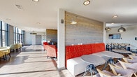 Located at 5104 Jacobs Rd in Seneca, SC, this single-floor community living space offers a generously sized, open-plan layout designed for comfort and social interaction. The space is defined by a striking combination of modern architecture and warm, inviting design elements. Horizontal wood-paneled accent walls provide a natural texture that contrasts beautifully with the crisp white walls and smooth, light-toned hardwood flooring found throughout.

A dramatic wall of floor-to-ceiling windows runs along one side of the space, flooding the interior with abundant natural light and offering expansive views of the surrounding outdoor landscape. This sun-drenched corridor is thoughtfully furnished with clustered seating arrangements featuring patterned upholstered chairs and round tables, ideal for casual gatherings.

The main lounge area is anchored by a tufted sofa, vibrant coral accent chairs, and a rustic wooden coffee table, all illuminated by a stylish geometric chandelier. A bold, large-scale abstract artwork serves as a focal point near the window seating. Additional zones include a dining area with long communal tables and cross-back chairs, a cozy banquette with yellow and red upholstered bench seating, and intimate conversation nooks. Recessed and pendant lighting complement the natural light throughout, creating a warm and welcoming ambiance.