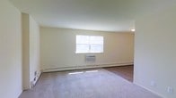 This single-floor space offers a clean and functional layout with a straightforward design. The main living area is finished with wall-to-wall carpeting that transitions to hard flooring near the entry, providing a comfortable and practical surface underfoot. Neutral, light-toned walls throughout the room create a bright and airy atmosphere, complemented by a large window that allows natural light to filter in and illuminate the space.

Climate comfort is well-addressed with both a wall-mounted window air conditioning unit and baseboard heating running along the perimeter of the room, ensuring year-round temperature control. A flush-mount ceiling light provides overhead illumination, keeping the space functional at all times of day.

The overall layout is open and unobstructed, offering flexibility for a variety of furniture arrangements. The minimalist design aesthetic keeps the focus on utility and livability, making it an easy space to personalize. The combination of natural light, dual climate control systems, and neutral finishes gives the space a practical yet inviting character suited for everyday living.