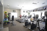This fitness center is a thoughtfully designed, single-floor facility offering a comprehensive workout environment for users of all fitness levels. The space is organized into distinct zones, each catering to different exercise needs.

The main cardio area features a row of Matrix treadmills and elliptical trainers, positioned to face large windows that flood the room with natural light and views of the surrounding exterior. Ceiling fans overhead ensure comfortable air circulation during intense workouts. Multiple flat-screen TVs are mounted along the walls, providing entertainment throughout the exercise session.

A dedicated strength training zone houses a cable machine system, a free-weight dumbbell rack, and a weight bench, offering a well-rounded selection of resistance equipment. Potted greenery is placed throughout the space, adding a refreshing, natural element to the environment.

A separate cycling studio, enclosed by floor-to-ceiling glass walls, contains Matrix stationary bikes and an elliptical, maintaining an open and airy feel while providing a distinct workout zone. A water station is conveniently located nearby.

An additional room, visible through the glass partition, features a colorful kids' activity area with bright furnishings, making the facility family-friendly. Neutral-toned carpet, recessed lighting, and a clean, modern aesthetic tie the entire space together cohesively.